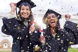 Two female, smiling students celebrating in caps and gowns outside on graduation day. They're throwing confetti, with diplomas in hand.