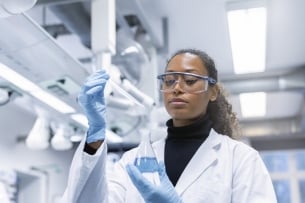A Black woman in a white lab coat and goggles holds a beaker of blue liquid.