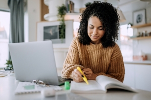 A woman holds a highlighter to a notebook while sitting in front of a laptop.