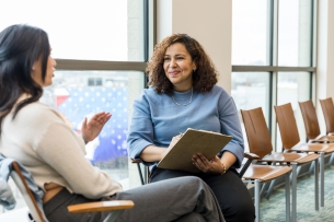 An adult female counselor smiles encouragingly as a young adult woman with her back to the camera gestures and shares.