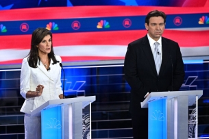 Nikki Haley, in a white suit, stands next to Ron DeSantis, who is wearing a dark suit and blue tie. Both are at podiums.