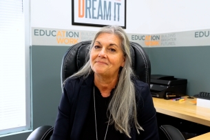 Jane Swift, a light-skinned woman with long gray hair wearing a black shirt, sits at her desk at the Education at Work headquarters in Arizona.
