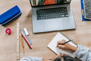 A student's hands can be seen taking notes in a notebook while a laptop is open to an online lesson in front of her. A collection of school supplies can be seen on the desk next to the laptop.