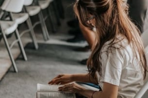 Close-up of a student sitting in a room reading a book with lines of chairs and a few people of different ages sitting and waiting