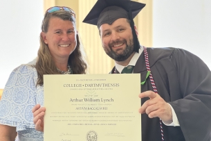 Photo of a light-skinned woman and a light-skinned man in academic regalia smiling while holding a diploma.