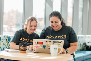 Two women wearing ADVANCE T-shirts sit at a table and smile at a laptop