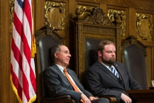 Two men sit in a state legislative chamber, flanked by American flags