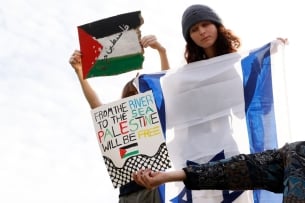 A pro-Israeli protester holds an Israeli flag unfurled. In front of her, hands can be seen holding pro-Palestinian signs, including one that reads “From the river to the sea, Palestine will be free.”