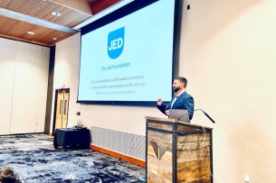 A man with a beard stands at a podium in front of a PowerPoint with the JED logo on it.