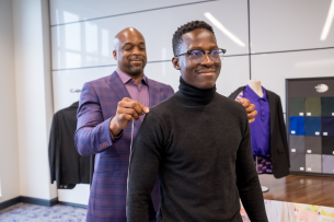 An older Black man, wearing a purple suit, measures a younger Black man's shoulders in a suit fitting.