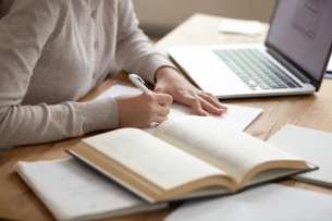 Student sits at desk taking notes from a book, computer open by her side