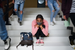 A Black student sits on the stairs looking distraught surrounded by peers.