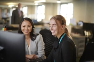 A supervisor trains a new hire on a computer. Both women are wearing headsets and professional clothing.