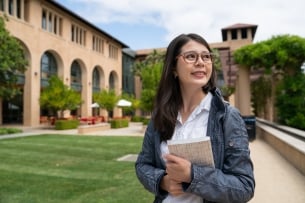 International student stands, holding a textbook, in front of a college building