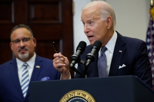 President Biden stands at a podium. A blurred-out Miguel Cardona is in the background.