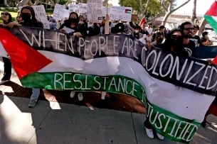 Student protesters have unfurled a large banner that features the Palestinian flag's colors and reads “When people are colonized, resistance is justified.” More protesters can be seen in the background, some holding signs that say things like “from the river to the sea, Palestine will be free” and “resistance against colonization is a human right.”