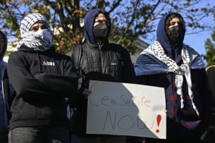 Three masked students attend a protest: two wear kaffiyehs. The student in the middle holds a sign that reads "Ceasefire Now!"