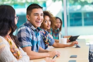 Male college student in a blue plaid shirt laughs with three peers at a desk.