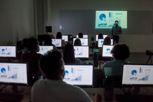 A group of students sits in front of computers, each showing charts and graphs on the screen. They are all facing toward the front of the room with their backs turned toward the camera.