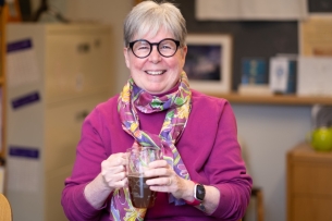 A smiling woman with gray hair and black glasses holding a large cup of tea