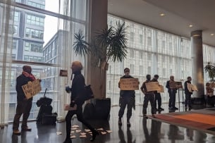 A photograph of seven demonstrators holding signs in a building as someone walks by. One sign says "Protect Pro-Palestine Activism" and another says "Stop Silencing Activism for Palestinians."