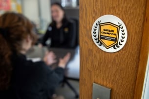 A sticker on the door to an office is in the foreground, with two people talking around a desk in the background. The sticker is white, with a yellow crest and the words "Career Champion" printed in black