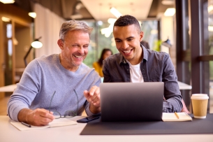 Mature businessman mentors younger colleague working on laptop at desk