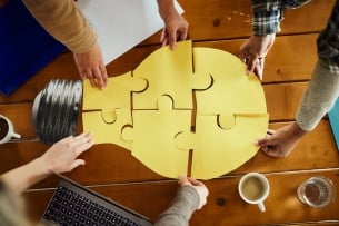 Various hands bring together puzzle pieces making up a bright light bulb