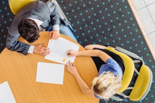 Aerial view of a student sitting at a desk next to a staff member with paperwork in front of them.