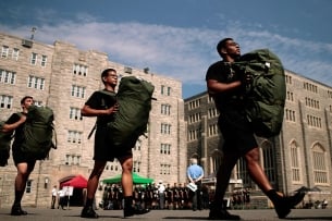Three students with heavy green packs walk past the facade of the U.S. Military Academy