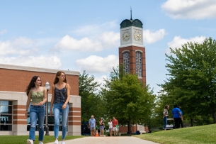 Two girls walk on a sidewalk with a college campus behind them.