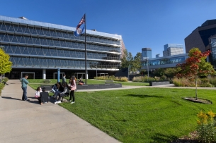 A group of people gather on a sidewalk next to a green lawn under a waving Minnesota state flag