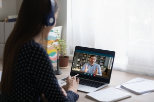 A woman wearing headphones faces a laptop. A man is on the screen of the laptop teaching.