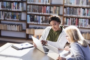 Two women working together in a library at a table. One has a laptop and the other is using a digital tablet to share information.