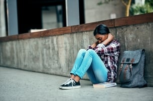 A female college student sits with her back to a wall and her head cradled in her hands, looking depressed, next to a backpack and book.