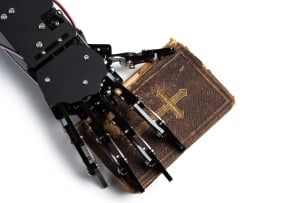 A robot hand is placed over a brown Bible with a cross on the cover.