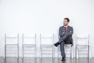 Man in suit sits alone impatiently waiting surrounded by a group of empty chairs