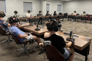 A group of students sit, all wearing virtual reality headsets. They are in a classroom and in a square formation with their desks. 