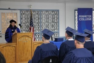 A group of students in caps and gowns sits in front of a professor at a podium. A blue “Perimeter College” banner is in the background.