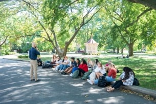 A Belmont University faculty member teaches a class outside to a row of students sitting on a curb with a green lawn and trees behind them.
