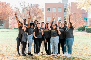 Students stand on a college campus in the fall wearing ADVANCE T-shirts