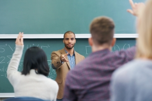 A young male teacher points at two students in the foreground who have their hands raised. Behind the instructor is a chalkboard with math equations