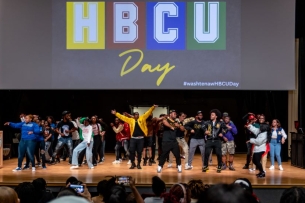 A group of students dance on a stage with a screen that reads “HBCU Day” in the background. 