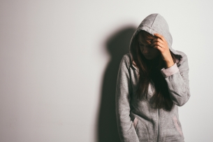 A college student stands in a poorly lit room with hood on her head and hand over her face, looking embarrassed.