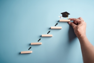 A series of ascending wooden blocks, arranged as steps, with upward arrows linking each of them, with a graduation cap atop the fifth and final step.