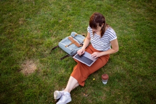 Woman sits on grass typing into a computer with a satchel and a paper cup of coffee on either side