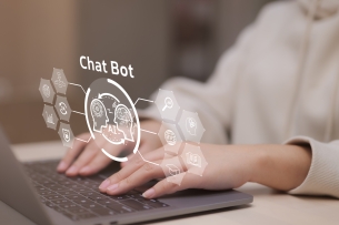 A close-up of a woman's hands resting on a laptop keyboard. An illustrated icon that reads "chat bot" hovers above the computer and the woman's hands.