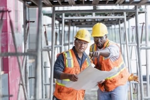 Two men in hard hats and high-visibility vests lean over a blueprint, one pointing.