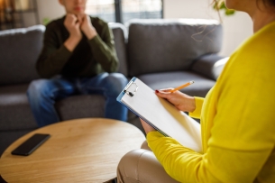 An over-the-shoulder photo of a female therapist meeting with a young male client. Neither person's face is visible.