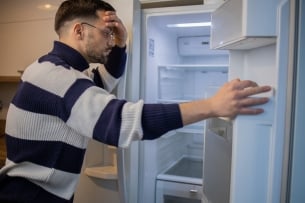 A young man holds a fridge door open and leans against the fridge, his head in his hand, as he surveys its contents: the fridge is empty.